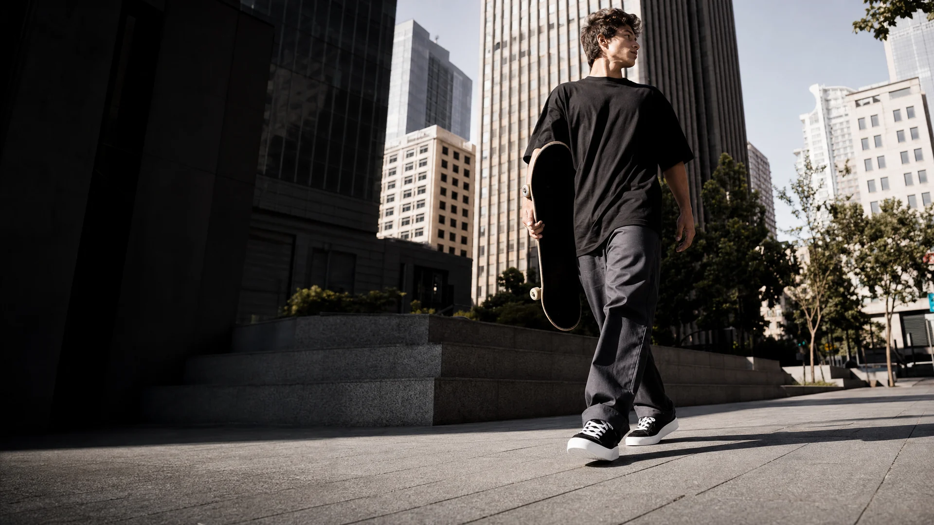 A person in black low-top trainers walking through a downtown plaza with a skateboard.